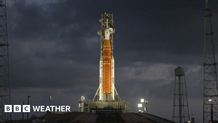 Orange rocket with white tip stands vertically ready to lift off lit up by flood lights