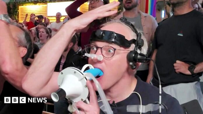 Prof Damian Bailey holds a white breathing mask connected to clear tubing up to James' face, who is seated and wearing a head-mounted device with wires and sensors attached. James has additional medical-style electrodes taped to the chest and arm