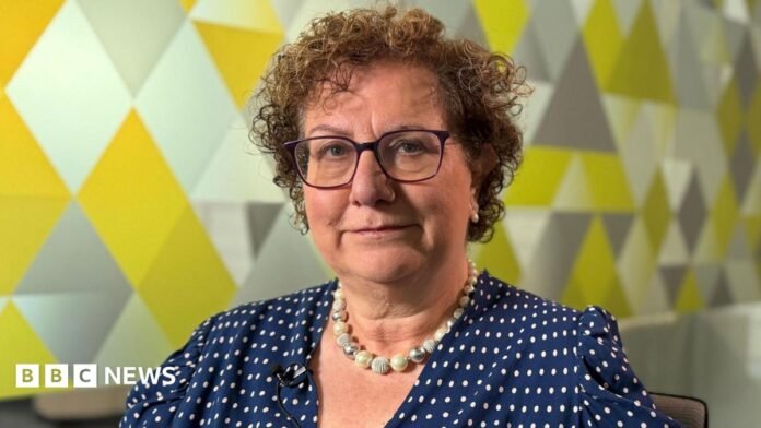 Dame Hilary Cass is sat looking at the camera in a meeting room. She is in front of a glass wall with a geometric pattern.