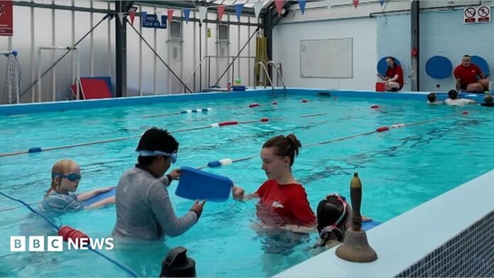 a swimming lesson in progress in a raised swimming pool. The pool is segregated into swimming lanes with a student being shown how to use a float by an instructor