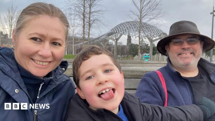 A selfie-style family photo of a woman and a man either side of a young boy, who is sticking his tongue out while smiling. They are all looking into the camera smiling.