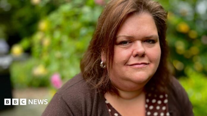 Caroline Fonjock looks at the camera. She is wearing a brown spotty dress and a chocolate brown matching cardigan. She has dark brown shoulder-length hair and silver hooped earrings on. She is standing in her garden with a yellow rose bush blurred in the background and some shrubbery to her right.