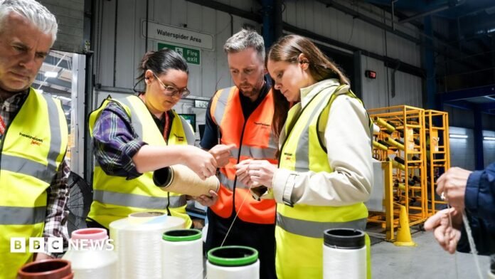 Four people wearing high-viz vests examine yarn from which parachute material is made, which is on big spools on a table. To the right a pair of hands can be seen while in the background is panelling and yellow shelves as well as a large door. 