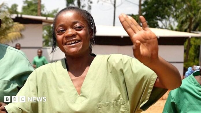 Health workers wearing green scrubs and white wellie boots dance and sing on a dusty ground