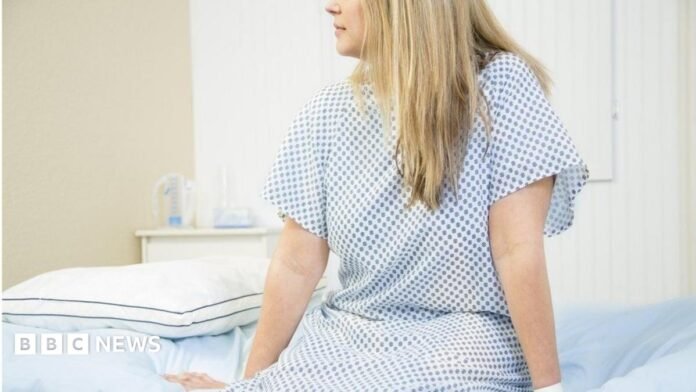 A woman wearing a white and blue hospital gown sits on the edge of a patient bed with her legs hanging down and her hands resting on the edge. She is looking to the left. The image is cut off above her nose. She has long, straight blonde hair.