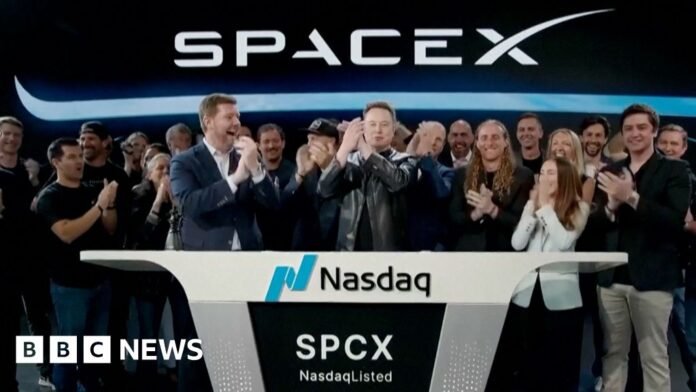 Elon Musk wearing a black leather jacket, stands in the center of crowds and in front of a SpaceX sign.