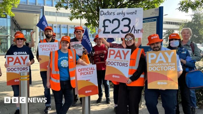 Resident doctors stand on a picket line outside Hull Royal Infirmary. They are holding orange and white signs featuring the BMA logo. The hospital building is visible in the background.