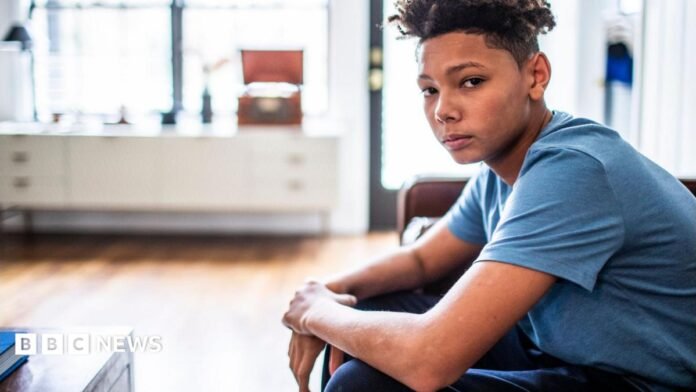 An anxious-looking teenager sits in a room, looking at the camera. 
