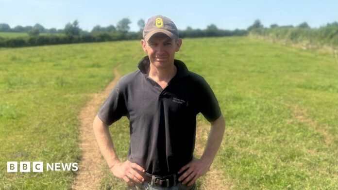 Rob wears a dark green polo shirt and a baseball cap and has his hands on his hips. He is standing in a grassy field with hedgerows and trees in the distance. 
