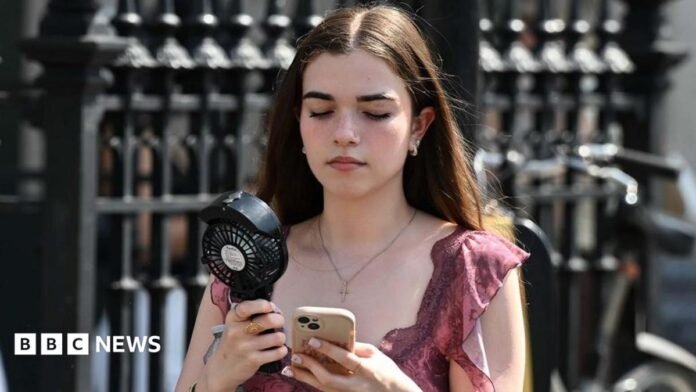 A young girl in a pink top with brown hair walks along a street carrying a portable black fan, whilst looking down at her phone in a clear case. 
