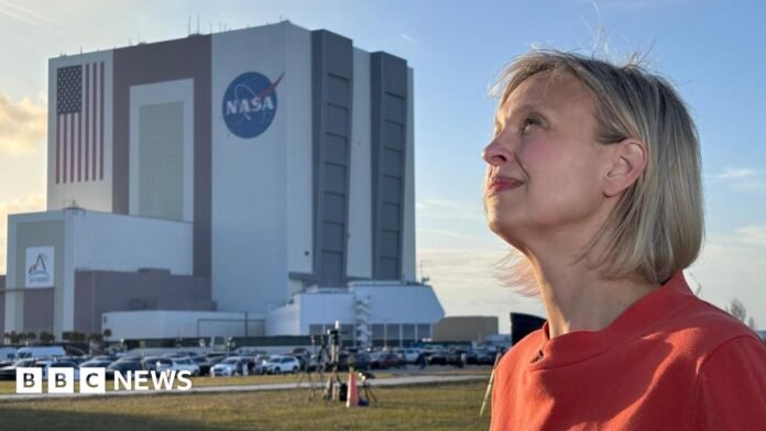 BBC Science Editor Rebecca Morelle stands about 100 metres in front of the cuboid Nasa mission control building. She is looking into the Sun as a shadow falls across the grass between her and the building. Some camera tripods are visible in the middle ground and a car park. Rebecca wears a red blouse or dress.