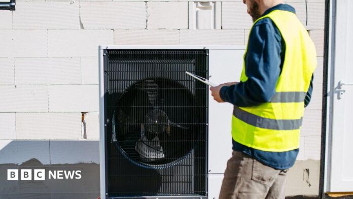 A black heat pump against a white breezeblock wall, a man stands to the right in brown jeans, blue shirt and a hi vis jacket holding a clipboard. He has a brown beard and the top of his head is cropped in the image
