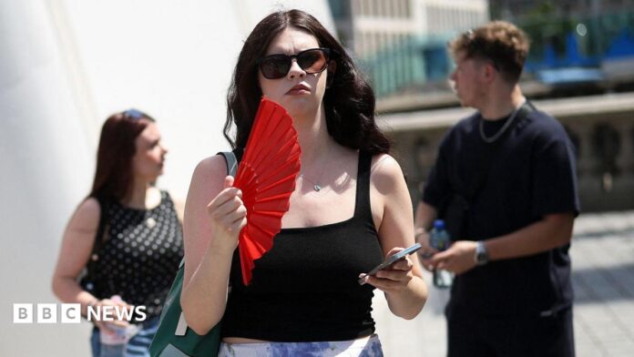 A woman on the street waves a red fan and holds her phone. She wears sunglasses and a black top.