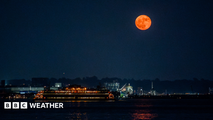 An orange-hued moon hangs in the sky over a river illuminated by lights from nearby properties