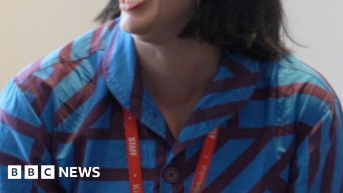 Woman with brown bob and glasses looks to her right and laughs, she is wearing a blue and red striped shirt and a red lanyard