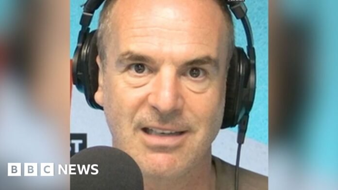 A man with short hair looks into the camera as he talks into a microphone