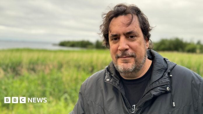 Dr. Eduardo Salazar-Ortuño. He has dark hair and grey beard, wearing a black raincoat and black t-shirt. He is standing on the shores of Lough Neagh.