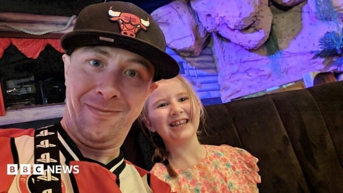 A man in a red and white football shirt and baseball cap smiles next to a younger girl in a pretty flowery dress