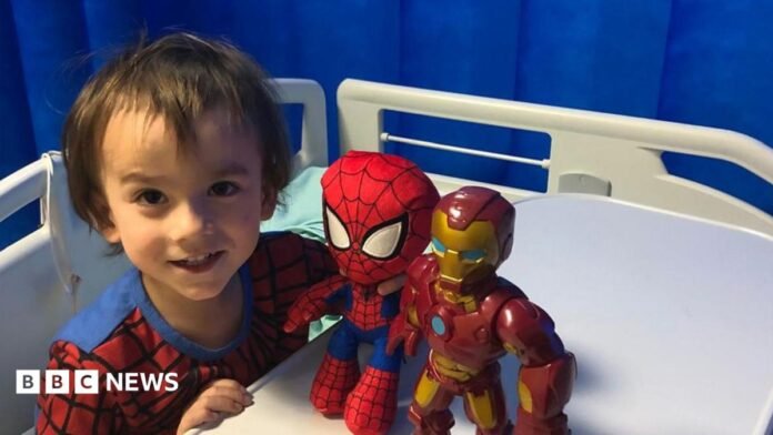 Theo Sebastian-Jenkin, then four-years old, sitting up on a hospital bed wearing a spider man costume while staring straight at the camera and playing with super hero toys.