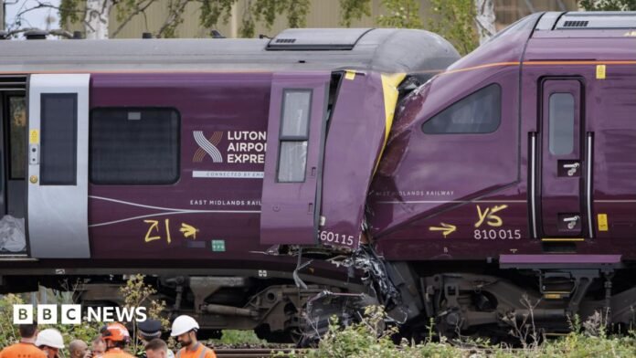 Two damaged trains, the front of one purple train is crushed into the end of another, with severe impact damage visible around the cab area.