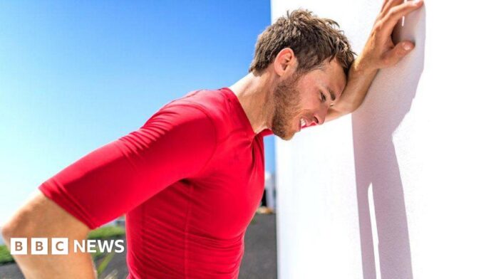 A sweating man with brown hair and wearing a red top leans against a white wall in the sun because he is overheating.