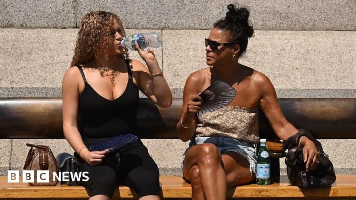 Two women sit on a bench in the sunshine. One woman is taking a sip from a bottle of water, while the other wears sunglasses and holds a fan. 