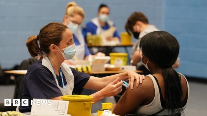 A nurse administers a vaccine does to a girl who is seated and faces away from the camera. The rest of the image is blurred but others can be seen also receiving their dose (file photo from March 2026)