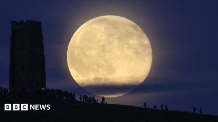 A Strawberry Moon rising above Glastonbury Tor in Somerset. The moon is large and bright, despite some cloud cover at the bottom of it. The silhouette of Glastonbury Tor can be seen to the left of the frame and there are people at the bottom of the frame. They are standing on the hill.