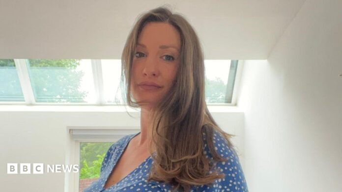A pregnant woman standing up in a room with white walls and a window behind her. She has long light brown hair and is wearing a blue dress and white dots.