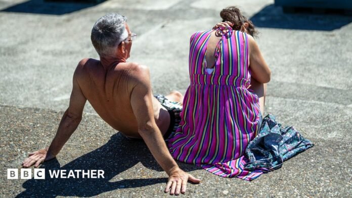 A man and a woman sit in the sun. The man is topless and leans back, the woman is in a purple sun dress has her head lowered and shields herself from the sun