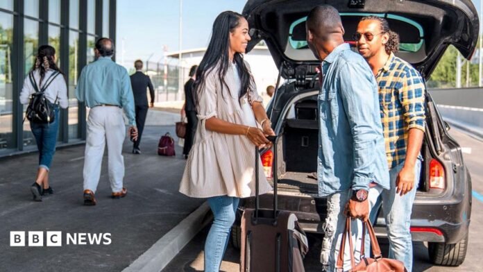 A sign outside Gatwick airport reads 'check-in & bag drop; north terminal Gatwick', while people walk past wheeling baggage along the concrete floor, image taken in 2024.
