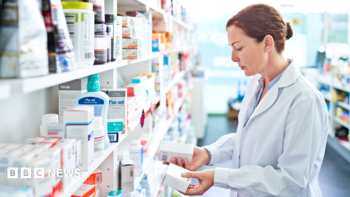 Pharmacist standing in front of shelves in pharmacy, holding medicine boxes in hands.