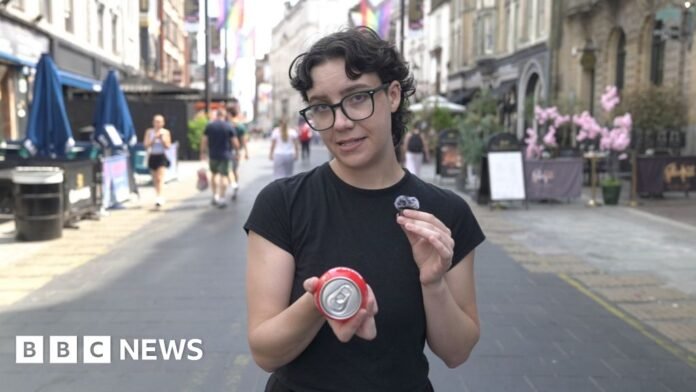A woman with black rimmed glasses in a black t shirt is holding a red spherical plastic and metal drink called a BuzzBall in her right hand. In her left hand is a bluetooth microphone. In the background there are stone buildings that house a mixture of shops, bars and restaurants, there is also the rainbow themed pride flag and multiple people walking around the centre of Cardiff. There are also dark blue parasols and bright pink flowers on either side of the street decorating two of the aforementioned bars.