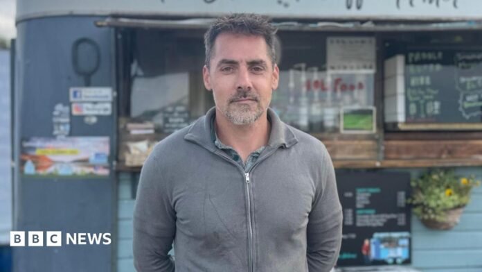 A man with grey hair is standing in front of a coffee cart. He is wearing a green shirt and a grey fleece with a zip at the front. 