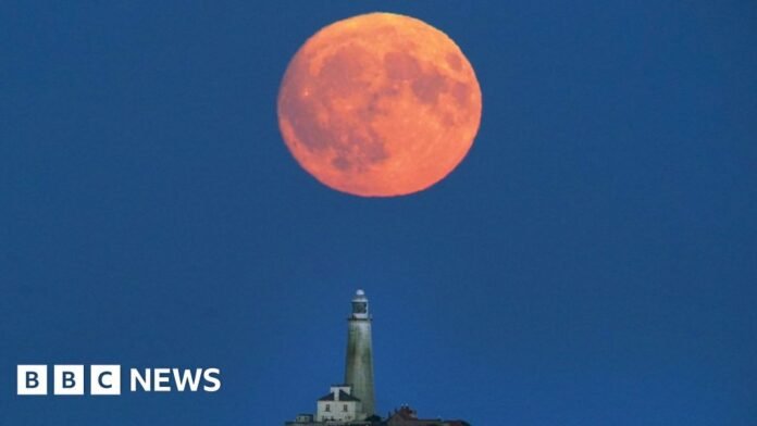 A red-orange full Moon hangs low in the night sky over a lighthouse