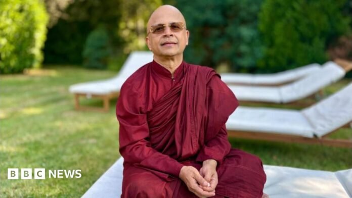 A bald man sits on a white lounger wearing dark red religious robes. He is in a garden and is wearing sunglasses.
