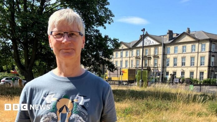 A man with white hair standing in a wildflower meadow. He is wearing glasses and a blue T-shirt with birds on it. Behind him stands a large, three-storey, Victorian-style building in yellow brick. 