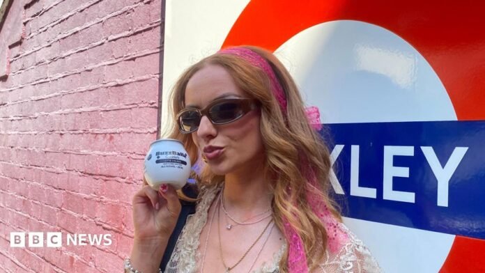 A young woman with long strawberry-blonde hair wears sunglasses and pouts at the camera, she is holding a white BuzzBallz container to her face. She is wearing sunglasses and a lace top with multiple necklaces on. Behind her is a London Transport Tube station sign and a pink brick wall.