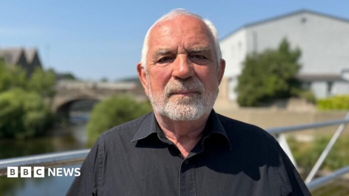 A man with grey hair and a grey beard in a black shirt stands on a bridge with a river, trees and a white building behind him. The background is blurred but there looks like another bridge over the water behind him.