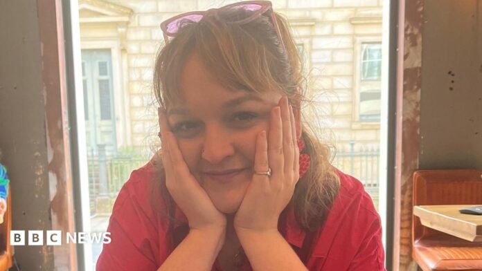 Daneka Etchells is pictured in a bright pink shirt sitting at a café table, resting her face in both hands and looking towards the camera. Large windows behind reveal a stone building outside.