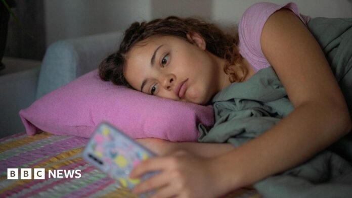 A stock image shows a young woman lying down with a glum expression as she scrolls on her smartphone