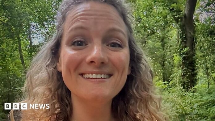 Zoe smiles at the camera in a selfie. She has light brown, curly hair and there are trees and woodland visible behind her. 