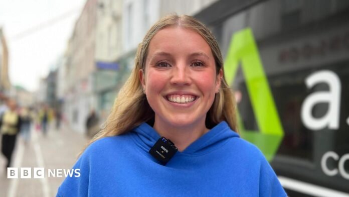 Eilidh is wearing a bright blue hoodie with the words “WHITE FOX” stands on a pedestrian shopping street. A small wireless microphone is clipped to the hoodie. Behind her, a shopfront window displays large white lettering and a green logo, while other pedestrians and buildings are visible in the softly blurred background.