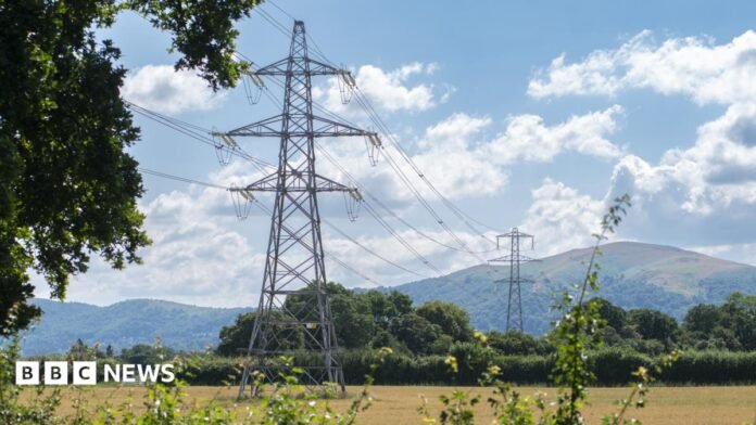Two pylons in a field, with rolling green hills in the background. Cables run across the field, over a hedgerow, to the second pylon in the background. 