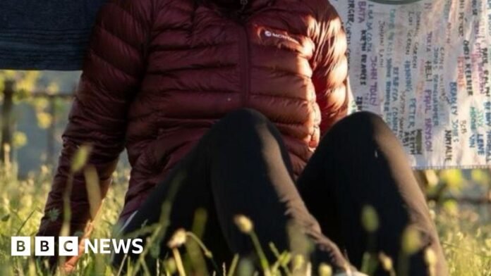 Kit sits in the grass outside. She wears a maroon down jacket and black trousers, has brown hair tied back in a low ponytail and there are blankets hanging on a wash line behind her.