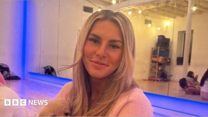 Ami Robertson sits in a Pilates studio. She is holding a dog on her lap and is smiling at the camera. She has long blonde hair.