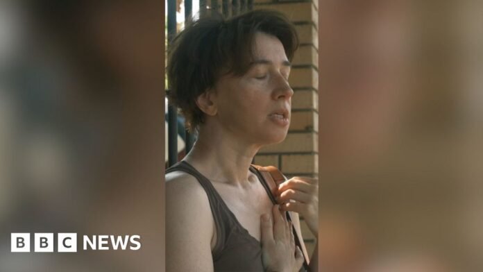 A woman with short hair stands in the street with her eyes closed and hand on her chest