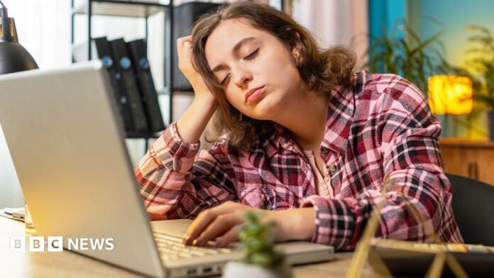 A woman with a brown bob, wearing a pink flannel shirt, sits at a table in front of an open laptop. She is resting on hand on the keyboard and the other is propping her head up. Her eyes are closed.