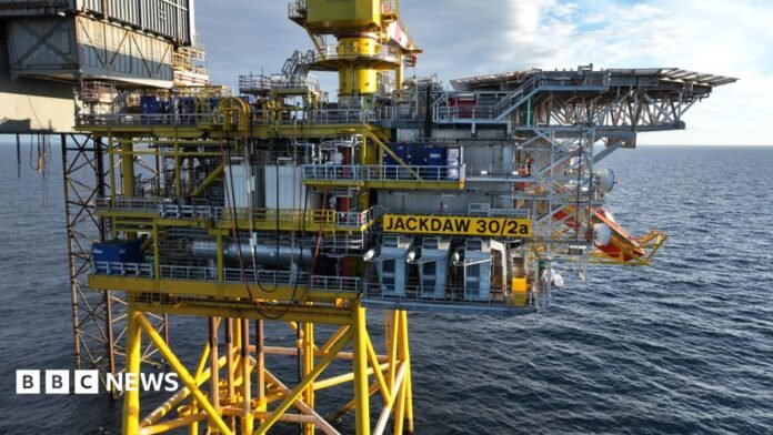 Offshore oil and gas platform marked “JACKDAW 30/2a” standing on yellow support legs above the sea, with industrial equipment, pipework and a crane visible under a partly cloudy sky.