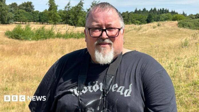 A man with short grey hair, a white goatee and black-rimmed glasses standing on a patch of grassland on a sunny day. Many trees are in the distance. He is wearing a black Motorhead t-shirt and has a pair of binoculars hanging around his neck.
